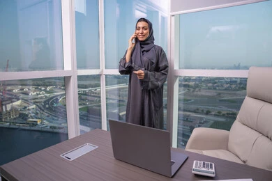 Remote work via mobile, enjoying exchanging conversations and news, an Arab Gulf Emirati woman wearing an abaya and hijab stands by the window making a phone call using her mobile phone, a comfortable work environment in Emirati companies.