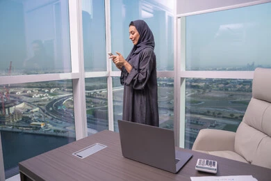 التواصل مع الأصدقاء عبر المنصات الإلكترونية، تصفح وسائل التواصل الاجتماعي باستخدام الجوال، تعابير وايماءات الفرح والسرور، امراة عربية خليجية اماراتية محجبة ترتدي العباءة و الحجاب تقف بجانب النافذة تمسك بيديها الهاتف المحمول التواصل مع الأصدقاء عبر المنصات الإلكترونية، تصفح وسائل التواصل الاجتماعي باستخدام الجوال، تعابير وايماءات الفرح والسرور، امراة عربية خليجية اماراتية محجبة ترتدي العباءة و الحجاب تقف بجانب النافذة تمسك بيديها الهاتف المحمول