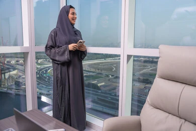 Browsing social media using a mobile phone, expressions and gestures of joy and happiness, chatting and communicating with friends online, looking at something, an Arab Gulf Emirati woman wearing a hijab and abaya standing by the window holding a mobile phone in her hands.