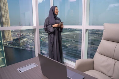 Expressions and gestures of joy and happiness, chatting and communicating with friends online, looking at something, browsing social media using a mobile phone, an Arab Gulf Emirati woman wearing a hijab and abaya standing by the window holding a mobile phone in her hands.