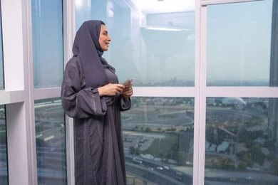 Chatting and communicating with friends online, looking at something, browsing social media using a mobile phone, an Arab Gulf Emirati woman wearing a hijab and abaya stands by the window holding a mobile phone in her hands, expressions and gestures of joy and happiness.
