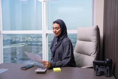 إعداد وثائق وتقارير العمل، التدقيق في قراءة الملاحظات، مراجعة ومتابعة الخطط والمشاريع المكتبية، امراه عربيه خليجيه اماراتية ترتدي العباءه و الحجاب تجلس على المكتب تمسك مجموعه من الاوراق البيضاء