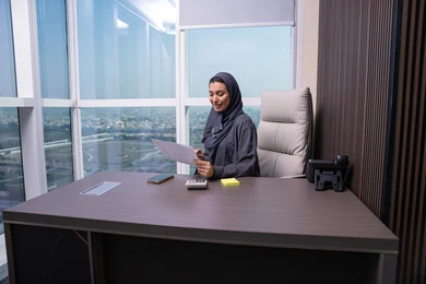 التدقيق في قراءة الملاحظات، اعداد التقارير الخاصة بالعمل، مراجعة ومتابعة الخطط والمشاريع المكتبية، امراه عربيه خليجيه اماراتية ترتدي العباءه و الحجاب تجلس على المكتب تمسك مجموعه من الاوراق البيضاء