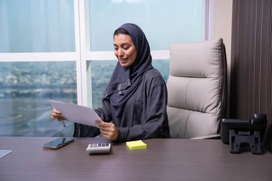 اعداد التقارير الخاصة بالعمل، مراجعة ومتابعة الخطط والمشاريع المكتبية، امراه عربيه خليجيه اماراتية ترتدي العباءه و الحجاب تجلس على المكتب تمسك مجموعه من الاوراق البيضاء، التدقيق في قراءة الملاحظات