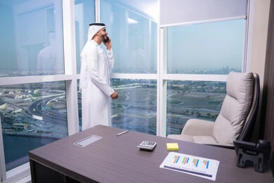 Making a voice call using a mobile phone, communicating and exchanging conversations and news with family and friends, an Arab Gulf Emirati man wearing a kandura and ghutrah stands by the window holding a mobile phone, using a modern and advanced technological device, professions and office work in the UAE.