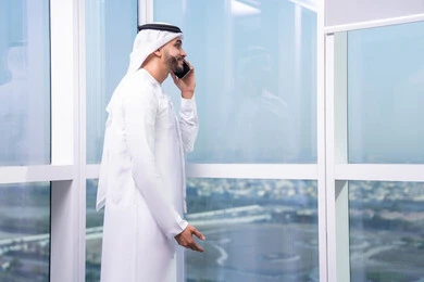 الاتصال عبر الجوال لإجراء مكالمة، التواصل وتبادل الأحاديث والأخبار مع الأهل والأصدقاء،  رجل عربي خليجي اماراتي يرتدي الكندورة و الغتره يقف بجانب النافذة يحمل الهاتف المحمول، استخدام جهاز تقني حديث ومتطور، مهن و أعمال مكتبية في الامارات