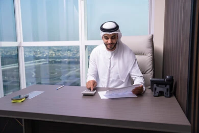 Reviewing the results extracted from the statistics, an Emirati Gulf Arab man wearing a kandura and ghutrah is working on the computer to complete tasks in the office, using a calculator to perform calculations, a clerical job in Gulf Arab companies.