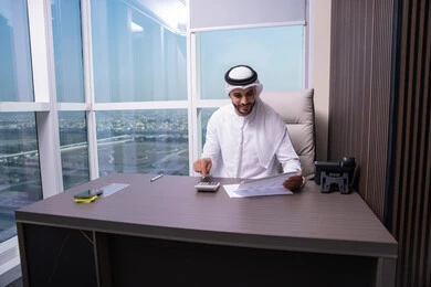 فحص البيانات الإحصائية للوصول إلى النتائج، رجل عربي خليجي اماراتي يرتدي الكندورة و الغتره يعمل على الحاسب لانجاز المهام في المكتب، استخدام الالة الحاسبة لاجراء العمليات الحسابية، وظيفة مكتبية في شركات الخليج العربي