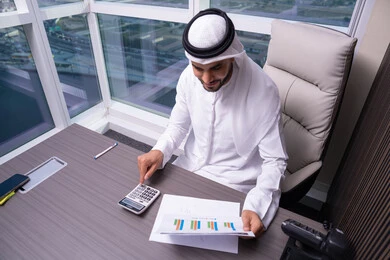Studying the results derived from statistical data, an Arab Gulf Emirati man wearing a kandura and ghutrah is working on a computer to complete tasks in the office, using a calculator to perform calculations, a clerical job in Gulf Arab companies.