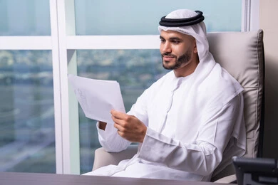 Preparing and following up on work reports, business feasibility studies, reviewing meeting results, an Arab Gulf Emirati man wearing a kandura and ghutrah sitting at the office holding a number of papers in his hand, browsing important notes.