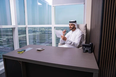 تنظيم مهام العمل وكتابة التقارير، دراسة جدوى الأعمال، مراجعة نتائج الاجتماع، رجل عربي خليجي اماراتي يرتدي الكندورة و الغتره يجلس فى المكتب يحمل بيده عدد من الأوراق، تصفح الملاحظات الهامة