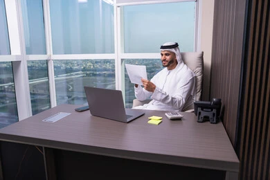 Preparation and review of performance reports, business feasibility studies, review of meeting results, an Arab Gulf Emirati man wearing a kandura and ghutrah sitting at the office in front of a laptop holding a number of papers in his hand, browsing important notes.