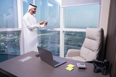 دراسة جدوى الأعمال، مراجعة نتائج الاجتماع، رجل عربي خليجي اماراتي يرتدي الكندورة و الغتره يقف فى المكتب  يحمل بيده عدد من الأوراق، تصفح الملاحظات الهامة، إعداد تقارير ومهام العمل