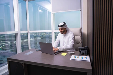إرسال إيميل إلكتروني من خلال الكمبيوتر، التواصل مع العملاء عن بعد، دمج التقنيات الحديثة و المتطورة مع العمل, مهن و أعمال مكتبية إدارية, رجل  عربي خليجي اماراتي يرتدي الكندورة و الغتره يجلس داخل المكتب و يقوم بالكتابة على لوحة المفاتيح