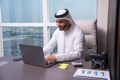 Using a laptop to send emails, communicate with clients remotely, integrating modern and advanced technologies with work, professions and administrative office work, an Arab Gulf Emirati man wearing a kandura and ghutrah sits in the office and types on the keyboard.