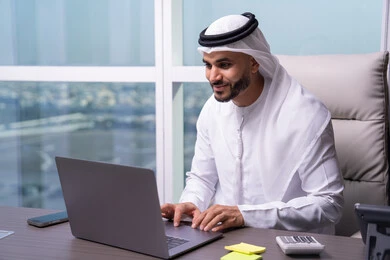 التواصل عبر البريد الإلكتروني من الكمبيوتر، التواصل مع العملاء عن بعد، دمج التقنيات الحديثة و المتطورة مع العمل, مهن و أعمال مكتبية إدارية, رجل  عربي خليجي اماراتي يرتدي الكندورة و الغتره يجلس داخل المكتب و يقوم بالكتابة على لوحة المفاتيح