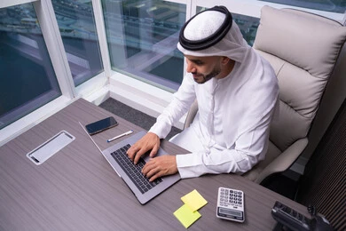إرسال بريد الكتروني عبر الحاسوب، التواصل مع العملاء عن بعد، دمج التقنيات الحديثة و المتطورة مع العمل, مهن و أعمال مكتبية إدارية, رجل  عربي خليجي اماراتي يرتدي الكندورة و الغتره يجلس داخل المكتب و يقوم بالكتابة على لوحة المفاتيح
