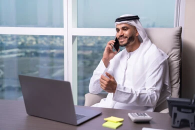 Project and task management remotely, using modern devices and advanced technologies, Emirati office professions and jobs, an Arab Gulf Emirati man wearing a kandura and ghutrah sitting at a desk in front of a laptop making a phone call via mobile.