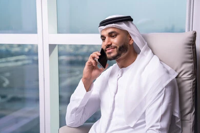 The concept of remote business management, the use of modern devices and advanced technologies, Emirati office professions and jobs, an Arab Gulf Emirati man wearing a kandura and ghutrah sitting at a desk making a phone call via mobile. The concept of remote business management, the use of modern devices and advanced technologies, Emirati office professions and jobs, an Arab Gulf Emirati man wearing a kandura and ghutrah sitting at a desk making a phone call via mobile.