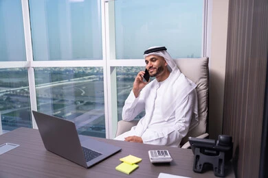 استخدام الأجهزة الحديثة والتقنيات المتطورة، مفهوم ادارة الاعمال عن بعد، مهن ووظائف مكتبيه اماراتية ، رجل عربي خليجي اماراتي يرتدي ا الكندورة و الغتره يجلس على المكتب امام الحاسب المحمول يقوم باجراء مكالمة هاتفية عبر الجوال
