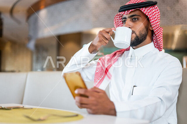 A Saudi Arabian Gulf man sitting in a café, enjoying drinking coffee, browsing the mobile phone, completing some work tasks in the café, working remotely, building relationships and friendships via the Internet, completing daily tasks, enjoying a calm atm