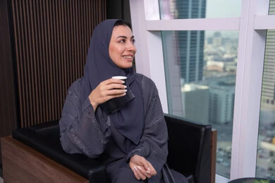 تعابير المتعة والاسترخاء، قضاء وقت الفراغ باحتساء المشروب المفضل, امراة  عربية سعودية اماراتية مبتسمة ترتدي العباءة و الحجاب تجلس على كرسي بجانب النافذة تقوم بشرب القهوة,  النظر الى شيء ما، ايماءات السعادة و السرور