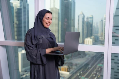 The use of modern and advanced devices and technologies, holding a business meeting online, a comfortable work environment in Emirati companies, an Arab Gulf Emirati woman wearing an abaya and hijab browsing social media news sites on a computer.