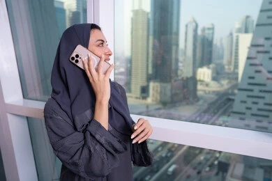 Communication and exchanging conversations and news with family and friends, a side image of an Emirati woman wearing an abaya and hijab standing by the window holding a mobile phone in her hand, using a modern and advanced technological device, making a phone call via the mobile device, professions and office work in the UAE.