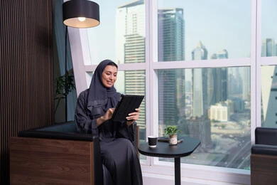 A profession and office job, conducting a video call, working remotely, accomplishing and managing tasks using a modern and advanced technical device, expressions of happiness and joy, an Arab Gulf Emirati woman wearing a hijab and abaya working on a tablet.