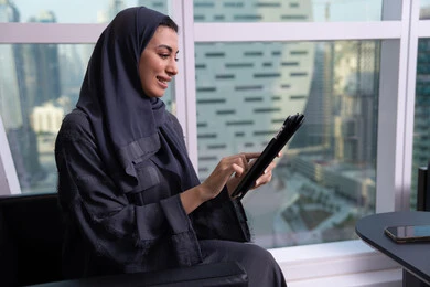 Visual communication via video, remote work, a profession and office job, accomplishing and managing tasks using a modern and advanced technical device, expressions of happiness and joy, an Arab Gulf Emirati woman wearing a hijab and abaya working on a tablet.