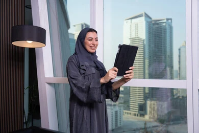 التواصل عبر مكالمة فيديو، العمل عن بعد، مهنة ووظيفة مكتبية، انجاز وادارة المهام باستخدام جهاز تقني حديث ومتطور, تعابير السعادة و السرور، امراة عربية خليجية اماراتية ترتدي الحجاب والعباءة تعمل على جهاز التابلت