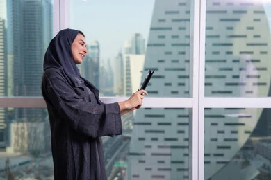 Making a video call, remote work, a profession and office job, accomplishing and managing tasks using a modern and advanced technical device, expressions of happiness and joy, an Arab Gulf Emirati woman wearing a hijab and abaya working on a tablet.