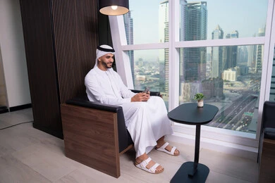 Emirati office professions and jobs, browsing social media, following global company news, an Arab Gulf Emirati man wearing a kandura and ghutrah sitting at his desk using a mobile phone and communicating with friends.