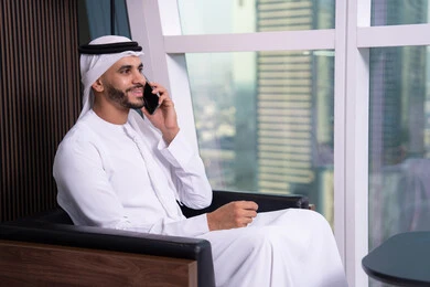 Phone communication using mobile, integrating technology and tech in practical life, remote work session, using advanced modern technologies, project study and reviewing the annual report, an Arab Gulf businessman from the UAE wearing a kandura and ghutrah sitting on a chair using a mobile phone.