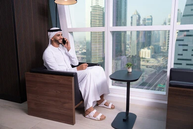 التواصل عبر الهاتف المحمول، دمج التقنية والتكنولوجيا في الحياة العملية، جلسه عمل عن بعد،  استخدام التقنيات الحديثة المتطورة، دراسه المشروع ومراجعه التقرير السنوي, رجل أعمال عربي خليجي اماراتي يرتدي الكندورة والغترة يجلس على كرسي يقوم باستخدام الجوال