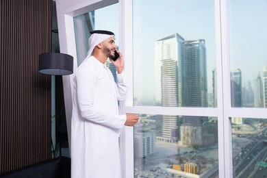 جلسه عمل عن بعد،  استخدام التقنيات الحديثة المتطورة، إجراء مكالمة صوتية عبر الهاتف المحمول، دراسه المشروع ومراجعه التقرير السنوي, رجل أعمال عربي خليجي اماراتي يرتدي الكندورة والغترة يقوم باستخدام الجوال، دمج التقنية والتكنولوجيا في الحياة العملية