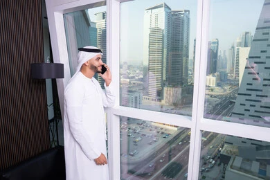 استخدام التقنيات الحديثة المتطورة، إجراء مكالمة صوتية عبر الهاتف المحمول، دراسه المشروع ومراجعه التقرير السنوي، جلسه عمل عن بعد, رجل أعمال عربي خليجي اماراتي يرتدي الكندورة والغترة يقوم باستخدام الجوال، دمج التقنية والتكنولوجيا في الحياة العملية