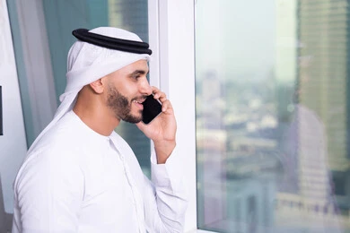 دراسه المشروع ومراجعه التقرير السنوي، إجراء مكالمة صوتية عبر الهاتف المحمول، استخدام التقنيات الحديثة المتطورة، جلسه عمل عن بعد, صورة مقربة لرجل أعمال عربي خليجي اماراتي يرتدي الكندورة والغترة يقوم باستخدام الجوال، دمج التقنية والتكنولوجيا في الحياة العملية