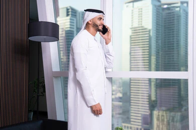 إجراء مكالمة صوتية عبر الهاتف المحمول، دراسه المشروع ومراجعه التقرير السنوي، استخدام التقنيات الحديثة المتطورة، جلسه عمل عن بعد, رجل أعمال عربي خليجي اماراتي يرتدي الكندورة والغترة يقوم باستخدام الجوال، دمج التقنية والتكنولوجيا في الحياة العملية