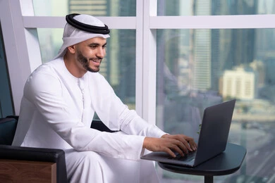 Using the keyboard to click, integrating work with technology and tech, managing and organizing business affairs through tech devices, an Arab Gulf Emirati man wearing a kandura and ghutrah sitting on a chair working on a laptop, administrative and office job professions.