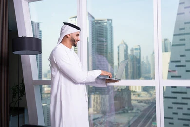 The use of a modern and advanced technical device, an administrative office profession and job, an Arab Gulf Emirati man wearing a kandura and ghutrah working on a laptop inside the office, companies and offices in the United Arab Emirates, managing and organizing business affairs, with the background of the Riyadh Towers.