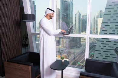 The use of a modern and advanced technical device, an administrative office profession and job, an Arab Gulf Emirati man wearing a kandura and ghutrah working on a laptop inside the office, companies and offices in the United Arab Emirates, managing and organizing business affairs, with the background of the Riyadh Towers.