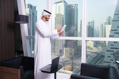 انجاز المهام وتحقيق الاهداف، دراسه وتتبع المشاريع، ملفات ووثائق خاصة بالعمل، الاحترافية والتفاني في الإنجاز، رجل عربي خليجي اماراتي يرتدي الكندورة والغترة يقف مقابل واجهة زجاجية مطلة على المدينة يقوم بقراءة الملاحظات، مهن ووظائف مكتبيه