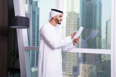 دراسه وتتبع المشاريع، ملفات ووثائق خاصة بالعمل، الاحترافية والتفاني في الإنجاز، رجل عربي خليجي اماراتي يرتدي الكندورة والغترة يقف مقابل واجهة زجاجية مطلة على المدينة يقوم بقراءة الملاحظات، انجاز المهام وتحقيق الاهداف، مهن ووظائف مكتبيه