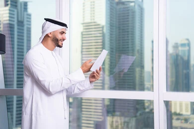 ملفات ووثائق خاصة بالعمل، الاحترافية والتفاني في الإنجاز، رجل عربي خليجي اماراتي يرتدي الكندورة والغترة يقف مقابل واجهة زجاجية مطلة على المدينة يقوم بقراءة الملاحظات، دراسه وتتبع المشاريع، انجاز المهام وتحقيق الاهداف، مهن ووظائف مكتبيه