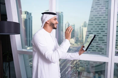 رفع الكف للأعلى بايماءات التحية والسلام، ايماءات السرور والسعادة, تصفح الانترنت واستخدام تقنيات حديثة, إجراء مكالمة فيديو عبر جهاز التابلت, العمل عن بعد من خلال الانترنت, رجل عربي خليجي اماراتي يرتدي الكندورة و الغتره البيضاء يقف مقابل واجهة زجاجية مطلة على المدينة يقوم بانجاز مهام العمل