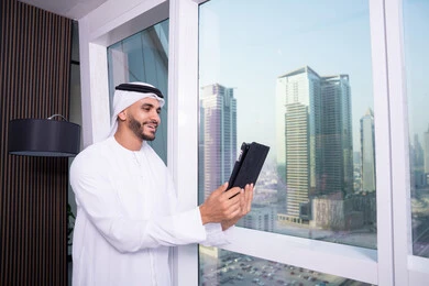 إجراء مكالمة فيديو عبر جهاز التابلت، ايماءات السرور والسعادة, تصفح الانترنت واستخدام تقنيات حديثة, العمل عن بعد من خلال الانترنت, رجل عربي خليجي اماراتي يرتدي الكندورة و الغتره البيضاء يقف مقابل واجهة زجاجية مطلة على المدينة يقوم بانجاز مهام العمل عبر الجهاز اللوحي