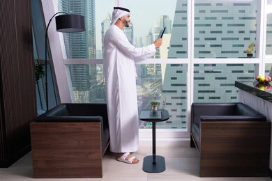 Remote work through the internet, gestures of joy and happiness, browsing the internet and using modern technologies, making a video call via a tablet, an Emirati Gulf Arab man wearing a kandura and a white ghutrah stands in front of a glass facade overlooking the city, completing work tasks via the tablet.