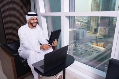 استخدام الإنترنت لإجراء مكالمة فيديو، ايماءات السعادة و السرور، مفهوم إدارة المشاريع عن بعد، رجل عربي خليجي اماراتي يرتدي الكندورة والغترة يجلس مقابل واجهة زجاجية مطلة على المدينة يقوم بانجاز مهام العمل عبر جهاز التابلت