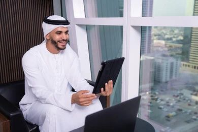 ايماءات السعادة و السرور، اجراء مكالمة فيديو عبر الانترنت، مفهوم إدارة المشاريع عن بعد، رجل عربي خليجي اماراتي يرتدي الكندورة والغترة يجلس مقابل واجهة زجاجية مطلة على المدينة يقوم بانجاز مهام العمل عبر جهاز التابلت